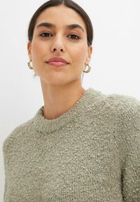 Maglione verde chiaro texturizzato con scollatura rotonda. Il tessuto presenta una superficie morbida e irregolare. Orecchini a cerchio dorati completano il look.