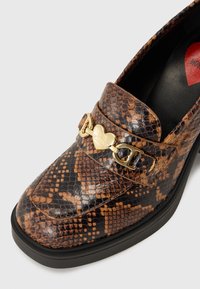 Καφέ και μαύρες ψεύτικες λωρίδες από φίδι loafers με ένα χρυσό λογότυπο καρδιάς, λεία υφή και μαύρη ελαστική σόλα.