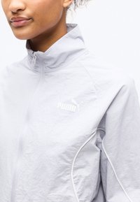 Κοντινό πλάνο ενός ατόμου που φοράει ανοιχτό γκρι ζακέτα Puma με φερμουάρ και ψηλό γιακά, με λευκό λογότυπο στο στήθος.