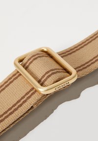 Sangle en tissu beige avec deux bandes marron passant à travers une boucle rectangulaire dorée gravée « COACH » sur un côté.