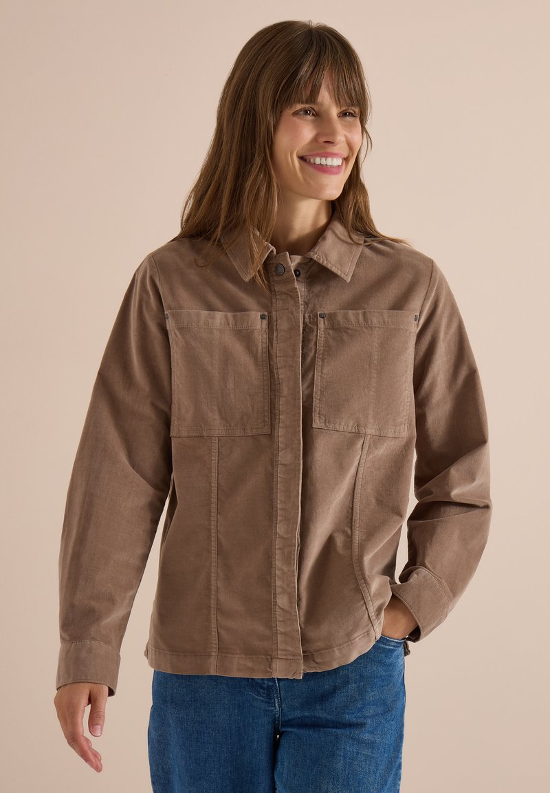 Femme souriante, portant une veste marron clair boutonnée avec de grandes poches avant et un jean bleu, debout devant un fond beige uni.
