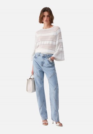 Giovane donna che indossa una maglia bianca in pizzo, jeans azzurri con dettagli curvi di paillettes, sandali bianchi con tacco, che tiene una borsa bianca.