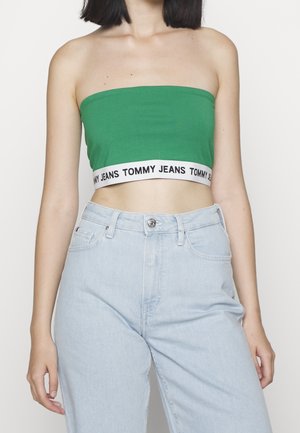 Frau trägt ein grünes Bandeau-Top mit weißem elastischem Band mit der Aufschrift "Tommy Jeans" und hellblaue, hochgeschnittene Jeans, gezeigt vom Hals bis zu den Hüften.