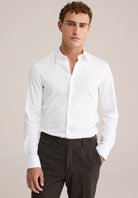 Jonge man in een witte long-sleeve casual overhemd en donkere broek, staand met één hand in zijn zak tegen een effen achtergrond.