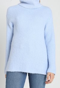 Ljusblå ribbad stickad turtlenecktröja bärs med blå jeans, sedd från nacken till höfterna med en hand avslappnat vid sidan.