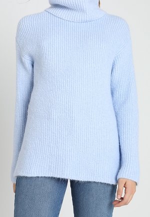 Světle modrý žebrovaný rolák, nošený s modrými džíny, zobrazený od krku po boky s jednou rukou uvolněně položenou po straně.