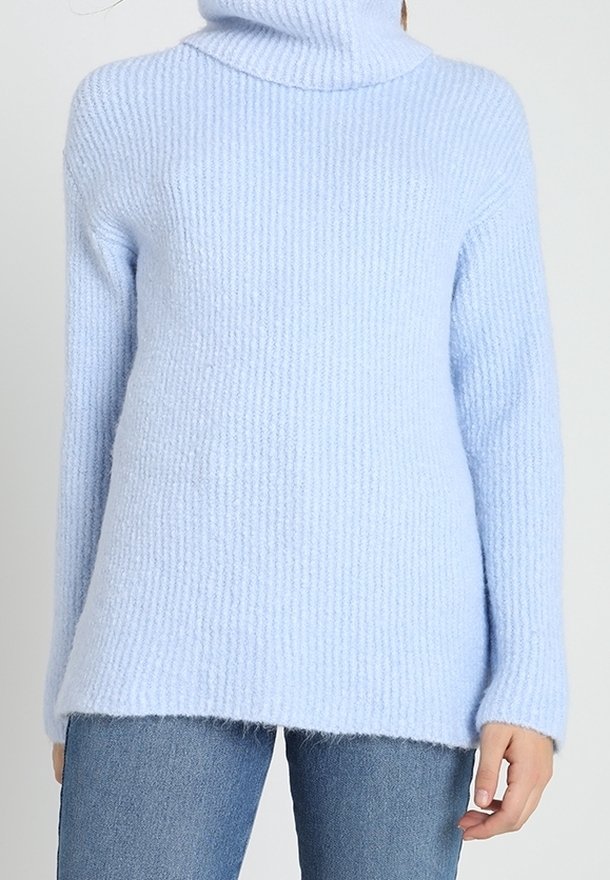 Ljusblå ribbad stickad turtlenecktröja bärs med blå jeans, sedd från nacken till höfterna med en hand avslappnat vid sidan.