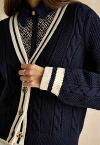 Tumši zils adīts kardigāns ar krēmkrāsas svītrām apkaklē un piedurkņu galos, ar lieliem pogām un uzlikts virs rakstainas krekla. Tekstūrēta adījuma dizains.