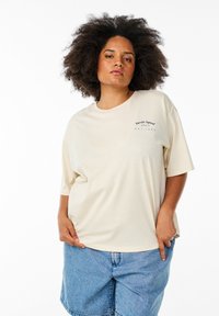 Beige oversized bomuld t-shirt med korte ærmer og rund halsudskæring. Forsiden har sort print: "Varsity Square | EST 1980."