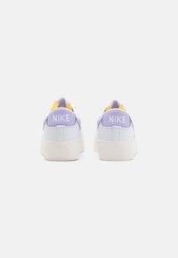 White sneakers with a padded purple heel, yellow accents, and the Nike logo prominently displayed on the heel tab. Flat sole design.