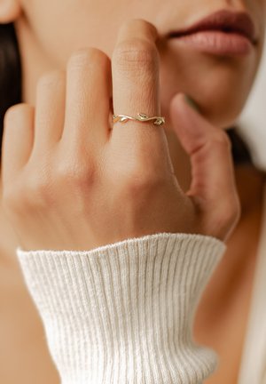 Anello d'oro con un design di vite attorcigliata, caratterizzato da piccoli accenti a forma di foglia. La mano è posizionata vicino a un maglione a coste di colore chiaro.