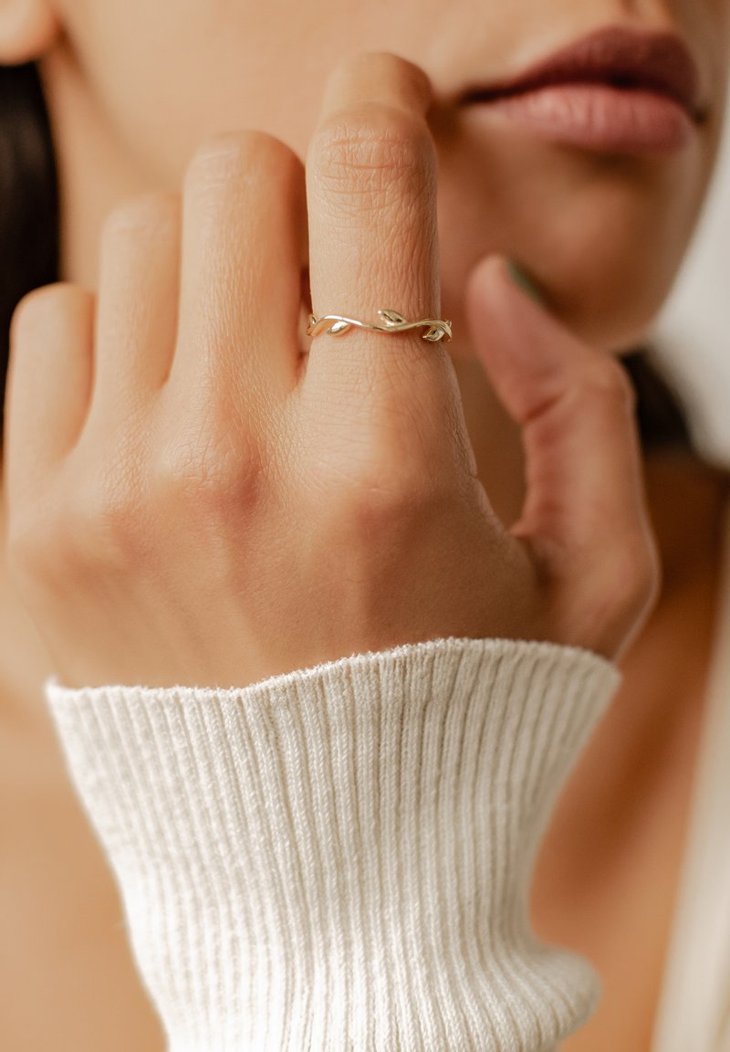Złoty pierścień z wirującym wzorem pnącza, z małymi akcentami liści. Dłoń znajduje się blisko swetra o prążkowanej, jasnej fakturze.