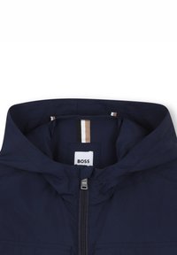 Giacca con cappuccio blu navy con zip frontale completa, caratterizzata da un'etichetta sul colletto, strisce a contrasto all'interno e tessuto liscio e leggero.