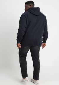 Marineblå hettegenser med en avslappet passform, med kengurulomme, ribbestrikkede ermer og kant. Kombinert med mørkegrå jeans og casual sko.