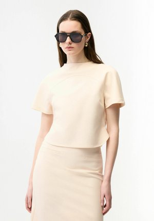 Kvinde iført beige kortærmet top og matchende nederdel, store sorte solbriller og guldørenringe mod hvid baggrund.