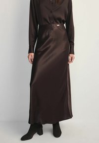 Kobieta ubrana w długą, ciemnobrązową satynową spódnicę, dopasowaną bluzkę z guzikami oraz czarne botki na obcasie, na jasnym tle.