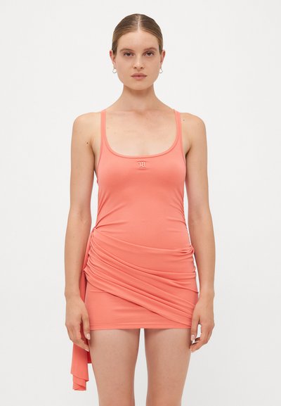 Femme avec les cheveux attachés, portant une mini-robe sans manches de couleur corail avec un détail drapé sur le côté et un petit logo sur la poitrine, se tenant devant un fond blanc.