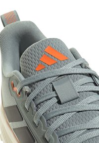 Zapato deportivo gris con tejido texturizado, acentos naranjas y diseño con cordones; cuenta con una lengüeta acolchada y suela sintética con ligera curvatura.