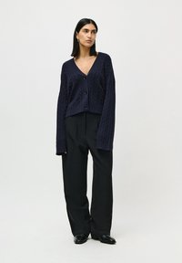 Kvinne som står med hendene ned langs siden, iført en marineblå kabelstrikket cardigan med knapper, svarte vide bukser og svarte sko, mot hvit bakgrunn.