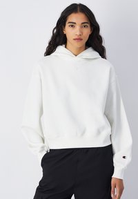 Sweatshirt à capuche blanc avec une coupe décontractée, des poignets côtelés et un logo sur la poitrine et la manche. Associé à un pantalon noir, le tout sur un fond clair.