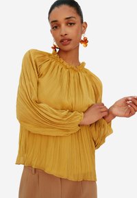 Blusa plissettata gialla senape con scollo a volant e maniche lunghe a palloncino. Tessuto leggero con una soffice e fluente texture.