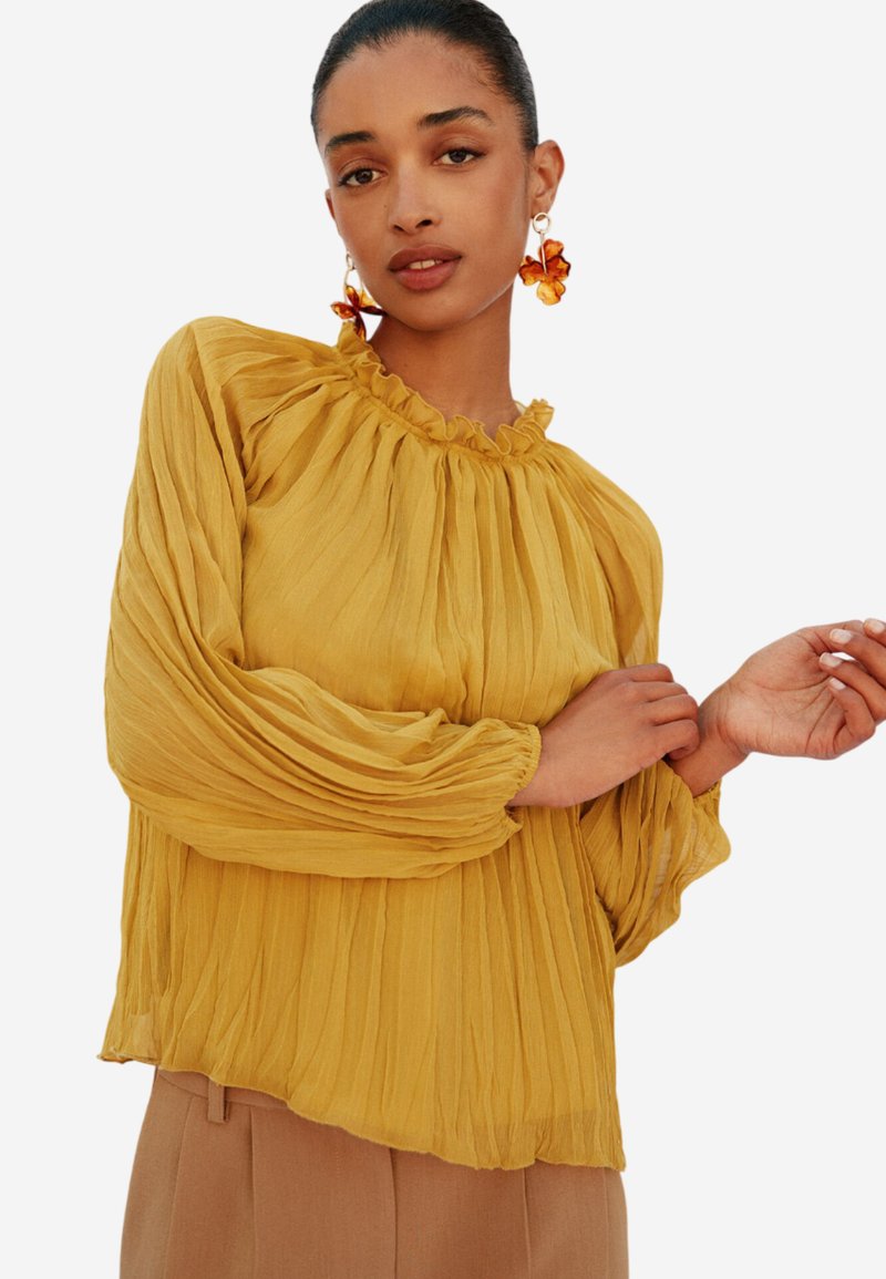 Blusa plissettata gialla senape con scollo a volant e maniche lunghe a palloncino. Tessuto leggero con una soffice e fluente texture.