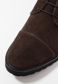 Bruine suède schoen met een ronde neus, zwarte rubberen zool en contrasterende stiksels. Veters zijn zichtbaar langs de bovenkant.