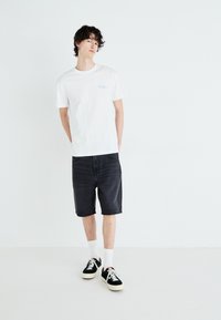 Weißes Baumwoll-T-Shirt mit blauem Logo, kombiniert mit schwarzen Jeansshorts. Das Model trägt weiße Socken und schwarze Sneakers mit roten Akzenten.