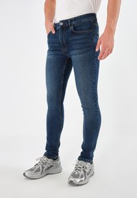 Vaqueros ajustados de color azul oscuro para hombre con una textura de vaquero suave, que cuentan con cinco bolsillos y un cierre de botón, combinados con zapatillas deportivas plateadas.