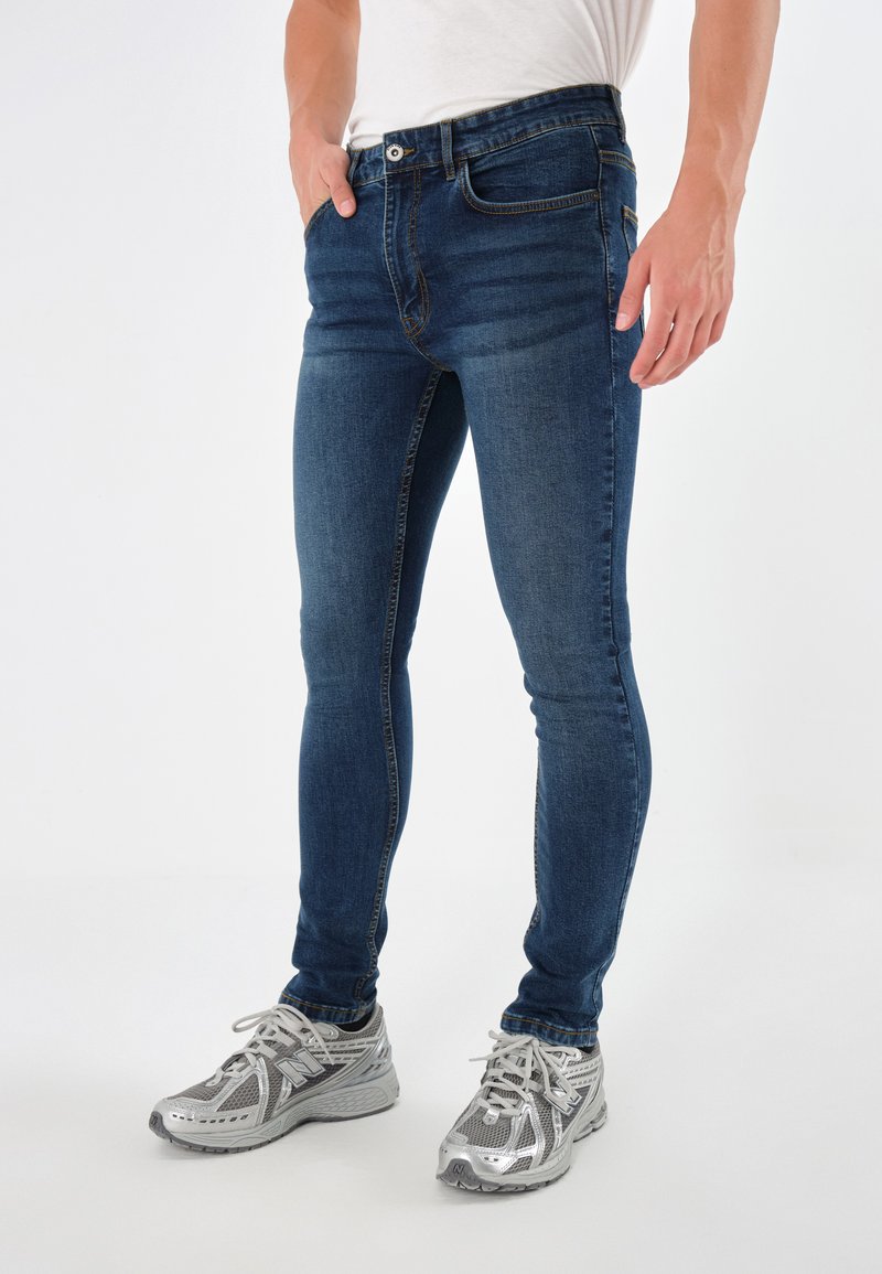 Vaqueros ajustados de color azul oscuro para hombre con una textura de vaquero suave, que cuentan con cinco bolsillos y un cierre de botón, combinados con zapatillas deportivas plateadas.