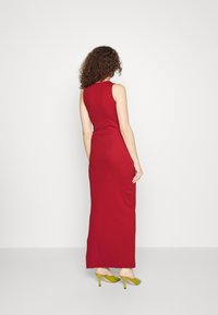 Rotes ärmelloses Maxikleid mit enganliegender Silhouette und glattem Material. Kombiniert mit grünen High-Heels. Neutrale Hintergrund.