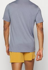 Hombre vistiendo una camiseta de manga corta gris claro y pantalones cortos amarillos mostaza, visto desde atrás con tatuajes visibles en ambos brazos.