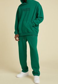 Groene hoodie met "GUESS ORIGINALS" gegraveerd op de borst, gecombineerd met bijpassende groene sweatpants en witte sneakers. Zacht katoenen materiaal.