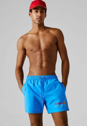 Giovane uomo con cappellino rosso e pantaloncini da bagno Red Bull Racing di un blu acceso, in piedi con le mani in tasca contro uno sfondo semplice.