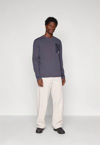 Langarm marineblauw shirt met een zakdetail, gecombineerd met licht beige broek. Model staat tegen een effen achtergrond en draagt zwarte schoenen.