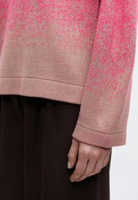 Μανίκι και τελείωμα μπεζ και ροζ πουαντελέ πλεκτού πουλόβερ με χέρι που ακουμπά χαλαρά δίπλα σε σκούρο καφέ παντελόνι, σε απλό φόντο.