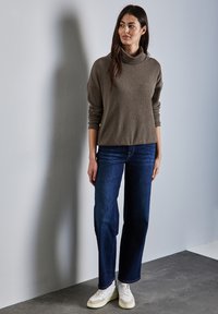 Högkragad, olivgrön tröja med vida ärmar, tillsammans med mörkblå raka jeans och vita sneakers med texturerade detaljer.