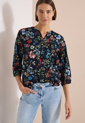 Femme portant un chemisier floral foncé à manches trois-quarts et un jean bleu clair, debout contre un mur beige et blanc.