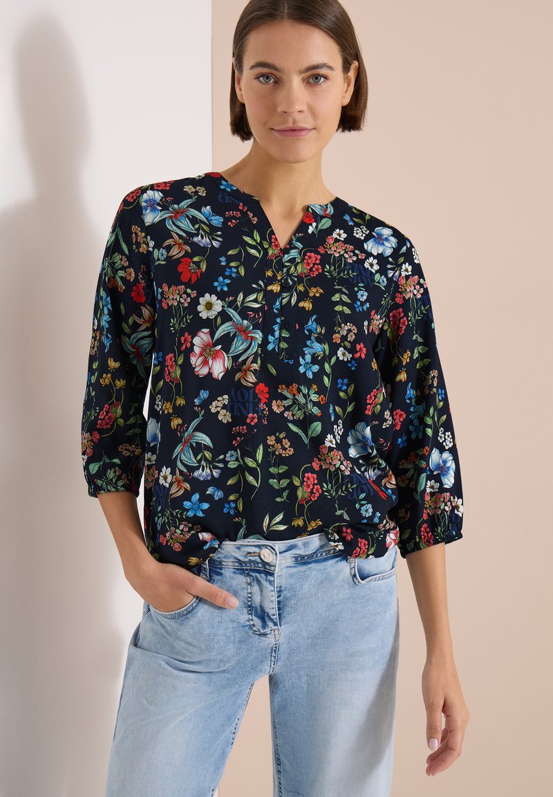 Femme portant un chemisier floral foncé à manches trois-quarts et un jean bleu clair, debout contre un mur beige et blanc.