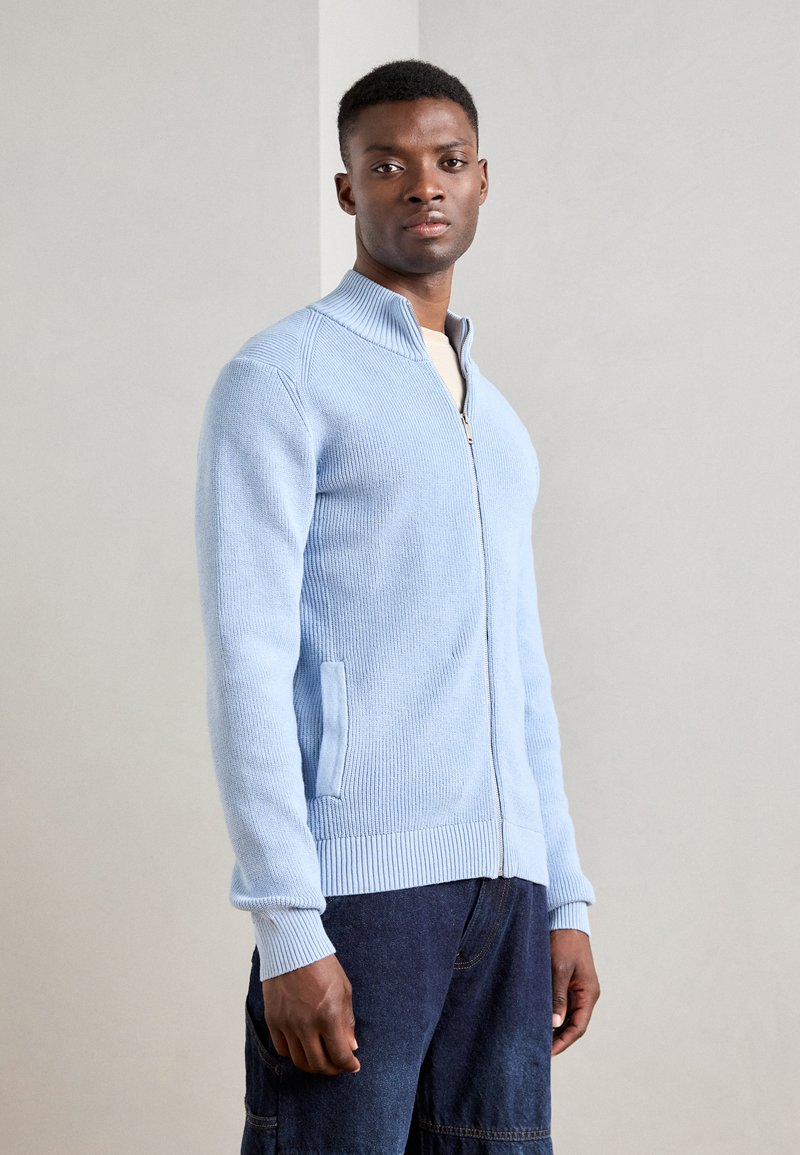 Light blue zip-up sweater with ribbed texture, standing collar, and side pockets, paired with dark denim pants.