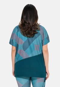 Blusa holgada color teal con patrones geométricos en azul claro, coral y azul oscuro. Textura suave, mangas cortas y diseño de dobladillo curvado.