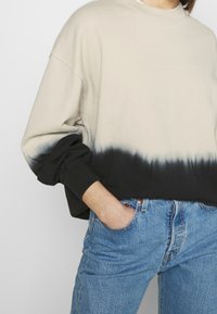 Felpa girocollo beige con sfumatura dip-dye nera. Maniche lunghe, vestibilità relaxed e texture morbida. Abbinata a jeans azzurri chiari.
