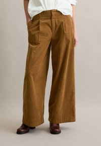 Bruine corduroy broek met wijde pijpen en voorplooien, voorzien van zijzakken en een gladde textuur, gecombineerd met donkerbruine enkellaarsjes.