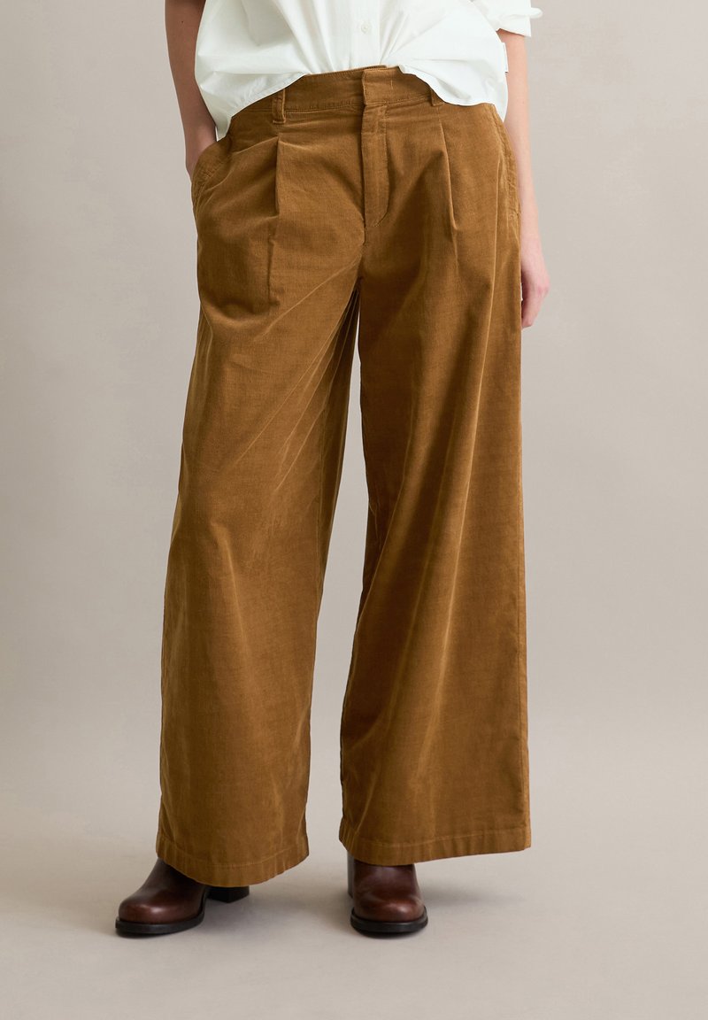 Bruine corduroy broek met wijde pijpen en voorplooien, voorzien van zijzakken en een gladde textuur, gecombineerd met donkerbruine enkellaarsjes.
