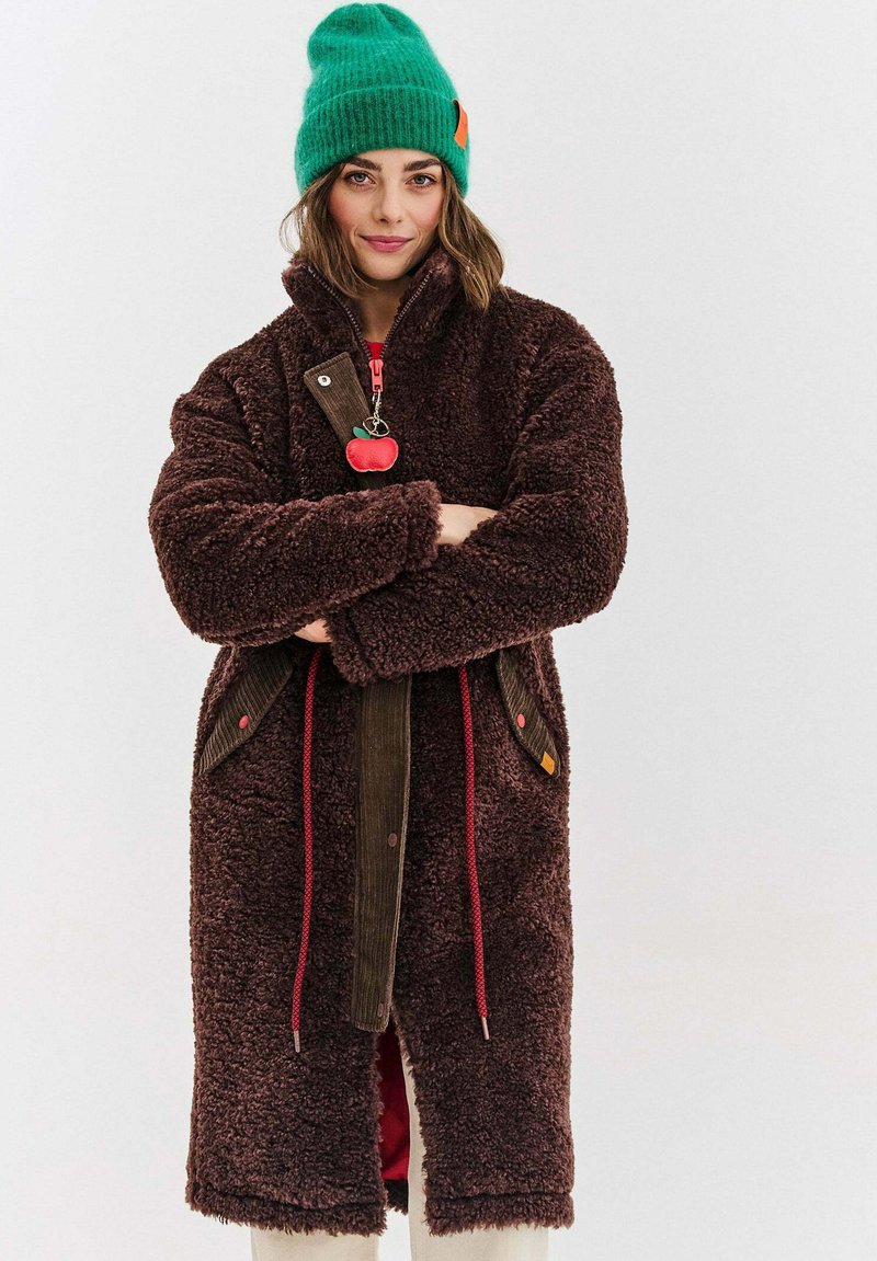 Mladá žena v tmavě hnědém texturovaném kabátu s červenými šňůrkami a zelenou pletenou čepicí, stojící s založenýma rukama před jednobarevným pozadím.