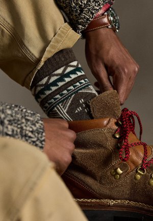 Brun ruskindsstøvle med røde snørebånd, tekstureret overdel og messing-øjer. Med et mønstret brunt og grønt sok, og opsmøgede beige bukser.