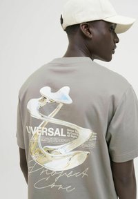 Pelēks īsām piedurknēm T-krekls ar grafisku druku, kas attēlo abstraktu dizainu un teksta detaļas aizmugurē; apvienots ar gaišu cepuri.