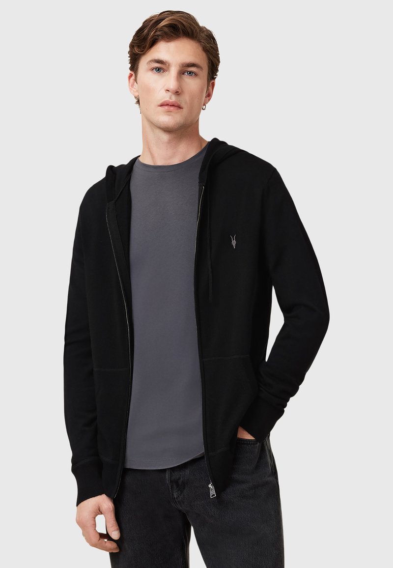 Sudadera negra con cremallera y bolsillos laterales, puños acanalados, textura suave, llevada sobre una camiseta gris de cuello redondo, con un sutil detalle de logo.