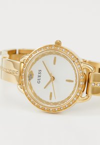 Gold wristwatch with a white dial, intricate bezel design, and crystal accents. Features a polished metal strap and minimalist hour markers.