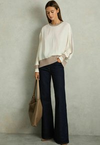 Krämfärgad tröja med vida ärmar, beige ribbad nederkant och manschetter, i kombination med mörka utsvängda jeans. Inkluderar en beige axelremsväska.